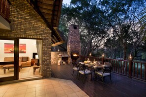 An outdoor deck with a brick fireplace, dining table, and chairs, surrounded by trees. - Kruger Park Lodge (Mbombela)