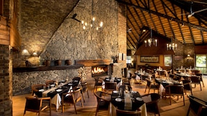A restaurant with a stone fireplace, wooden tables, and chairs arranged for dining. - Kruger Park Lodge (Mbombela)