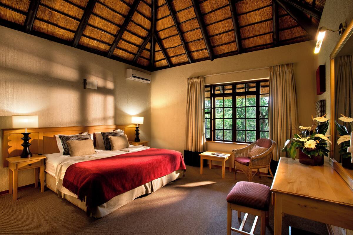 Une chambre d’hôtel avec un lit, un bureau, une chaise, une fenêtre avec des rideaux et un toit de chaume.
