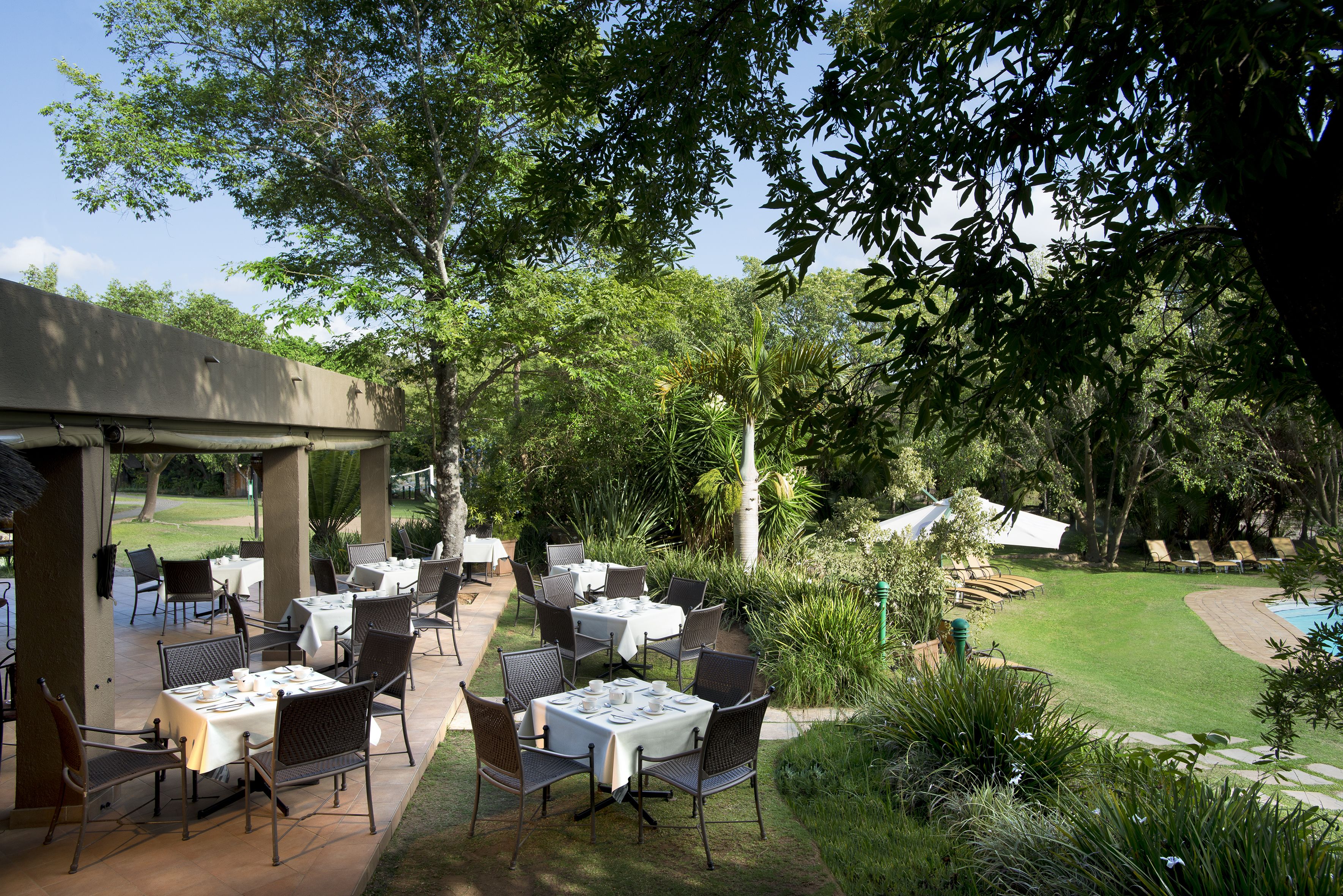 An outdoor patio with tables and chairs, surrounded by lush greenery and a pool.