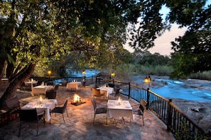 An outdoor dining area with tables set for two, surrounded by stone flooring, and overlooking a flowing river.