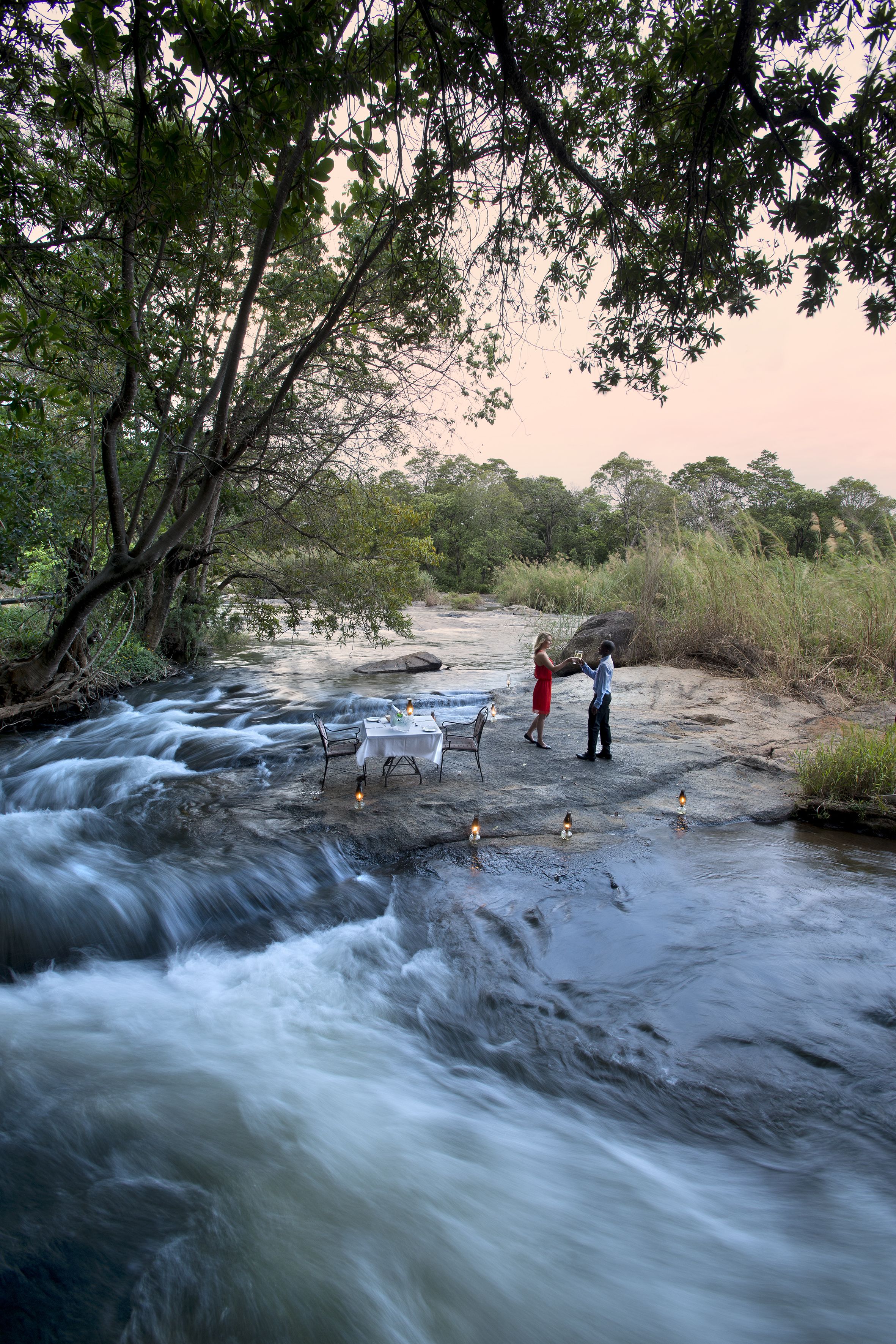 A river with a table and chairs set up on a rock, surrounded by trees.