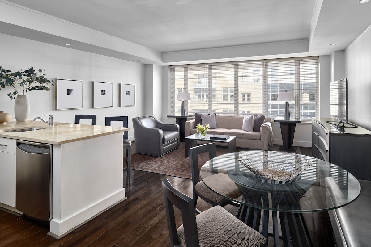 A modern hotel room with a kitchen area, a dining table with chairs, a sofa set, and large windows offering a city view.
