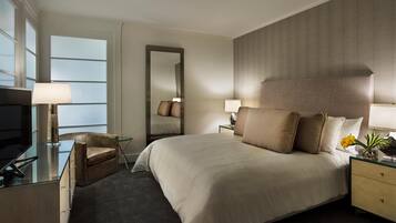 A hotel room with a large bed, a desk with a TV, a chair, a nightstand with a lamp, and a mirror.