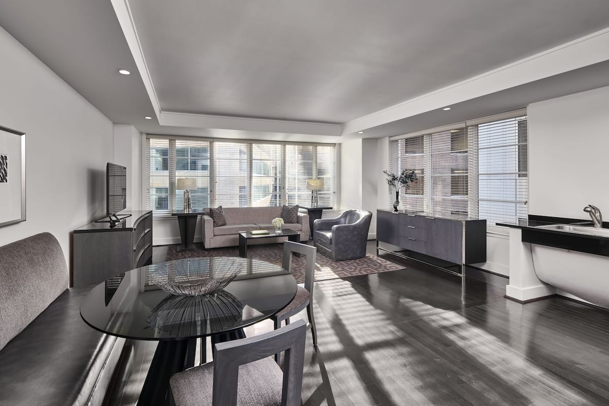 A modern living room with a glass dining table, a sofa, a chair, and a kitchen area with a sink and cabinets.