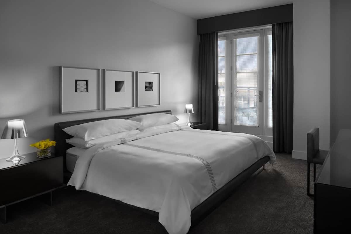 A modern hotel room with a large bed, a bedside table with a lamp, and a window with blinds.