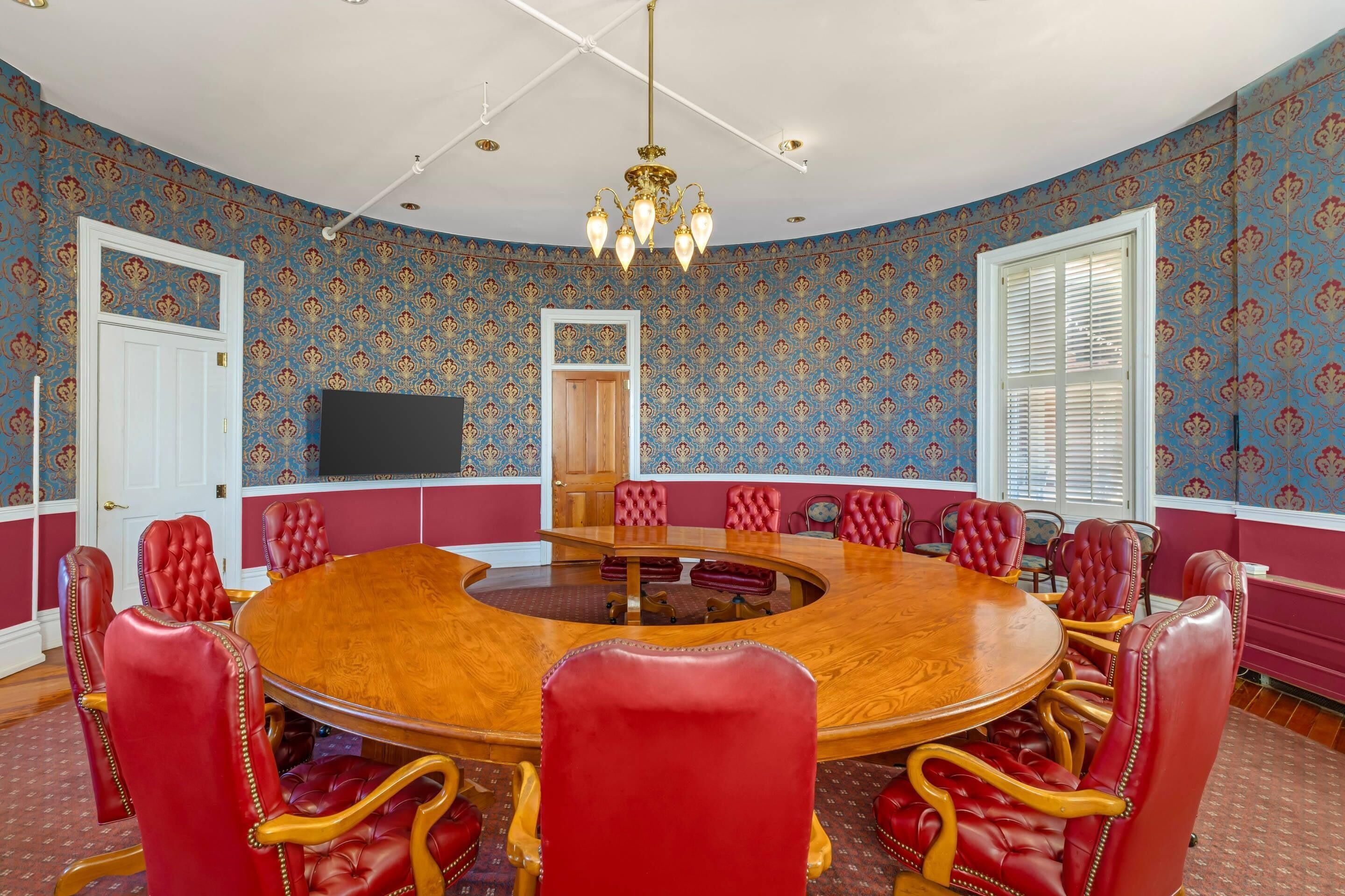 A conference room with a round wooden table, red leather chairs, a chandelier, patterned wallpaper, and a television on the wall.