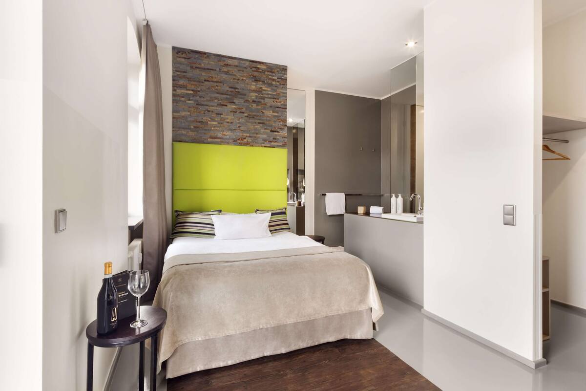 Una camera d'albergo moderna con un letto grande, una testiera a motivo di mattoni, una parete verde decorativa e un tavolino con una bottiglia e un bicchiere.