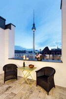 A balcony with two wicker chairs, a small table, and a vase of flowers. A bottle and glasses are on the table. The view includes a tower and buildings.