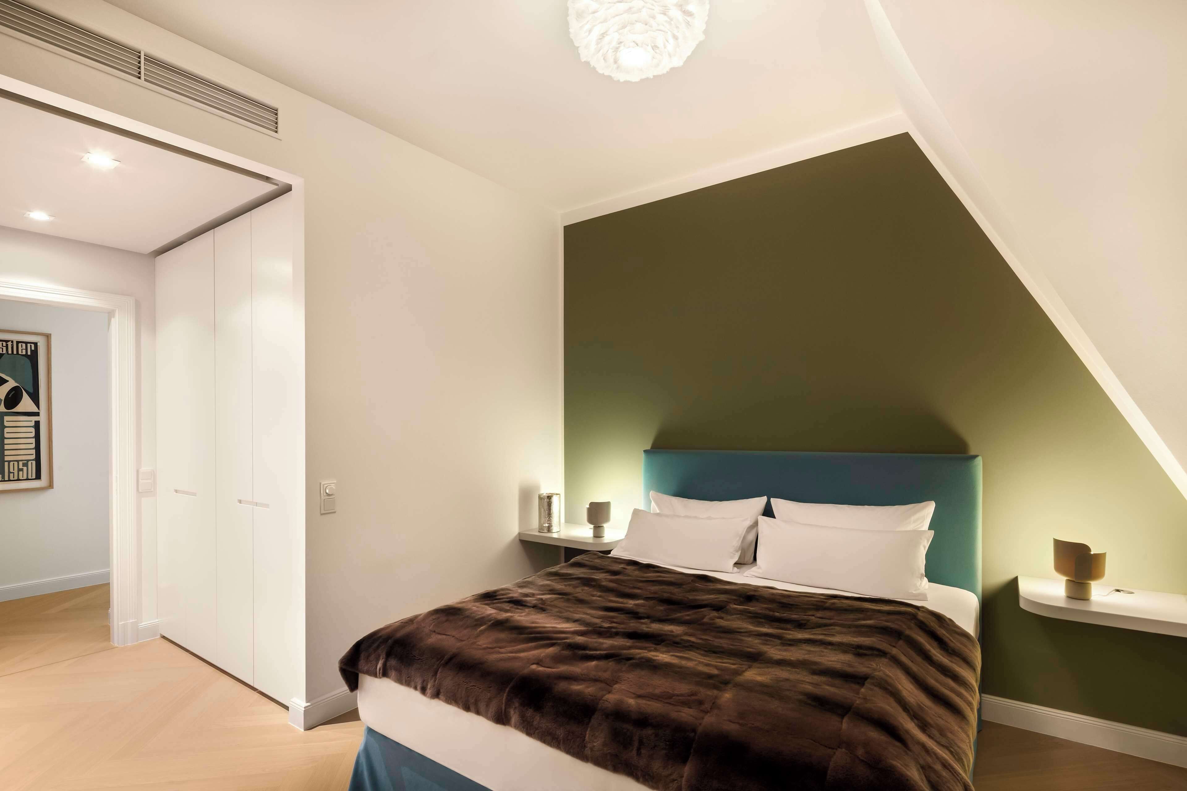 A bedroom with a bed, nightstands, a wall-mounted shelf, and a door leading to another room.