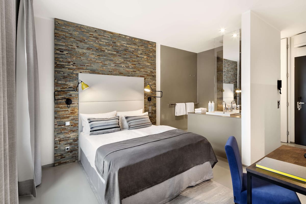 Una camera d'albergo moderna con una parete decorata con un motivo a mattoni, un letto grande con copripiumino grigio e bianco, un tavolino con una sedia, e un bagno visibile attraverso una porta aperta.