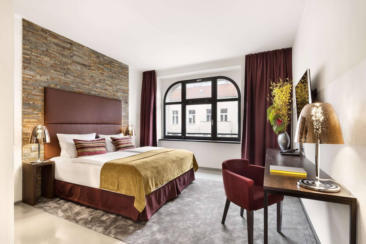 Una moderna camera d'hotel con un letto grande, una scrivania con una sedia, una finestra con tende e una lampada decorativa.