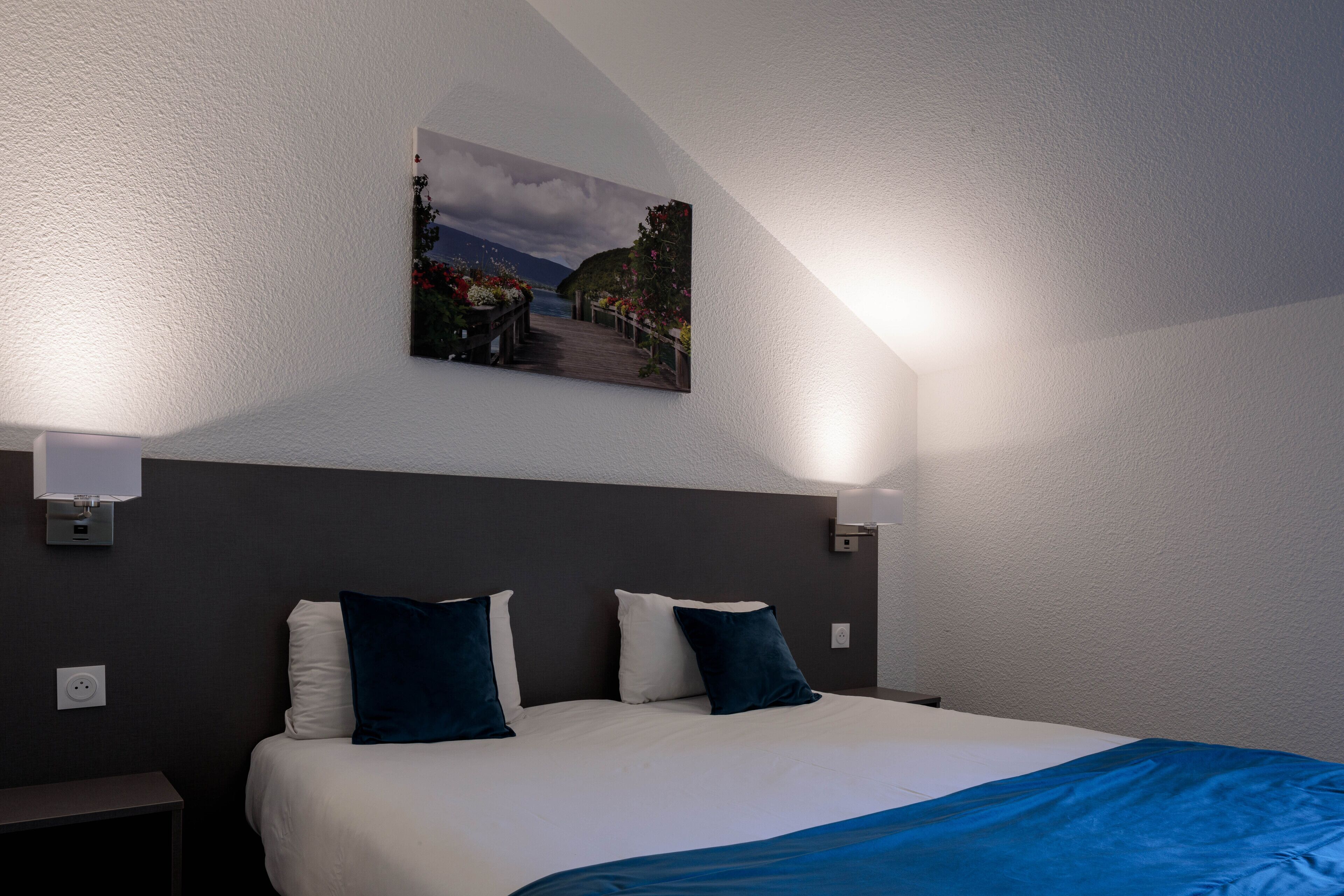 A bedroom with a bed, a nightstand, a wall-mounted lamp, and a painting of a landscape.