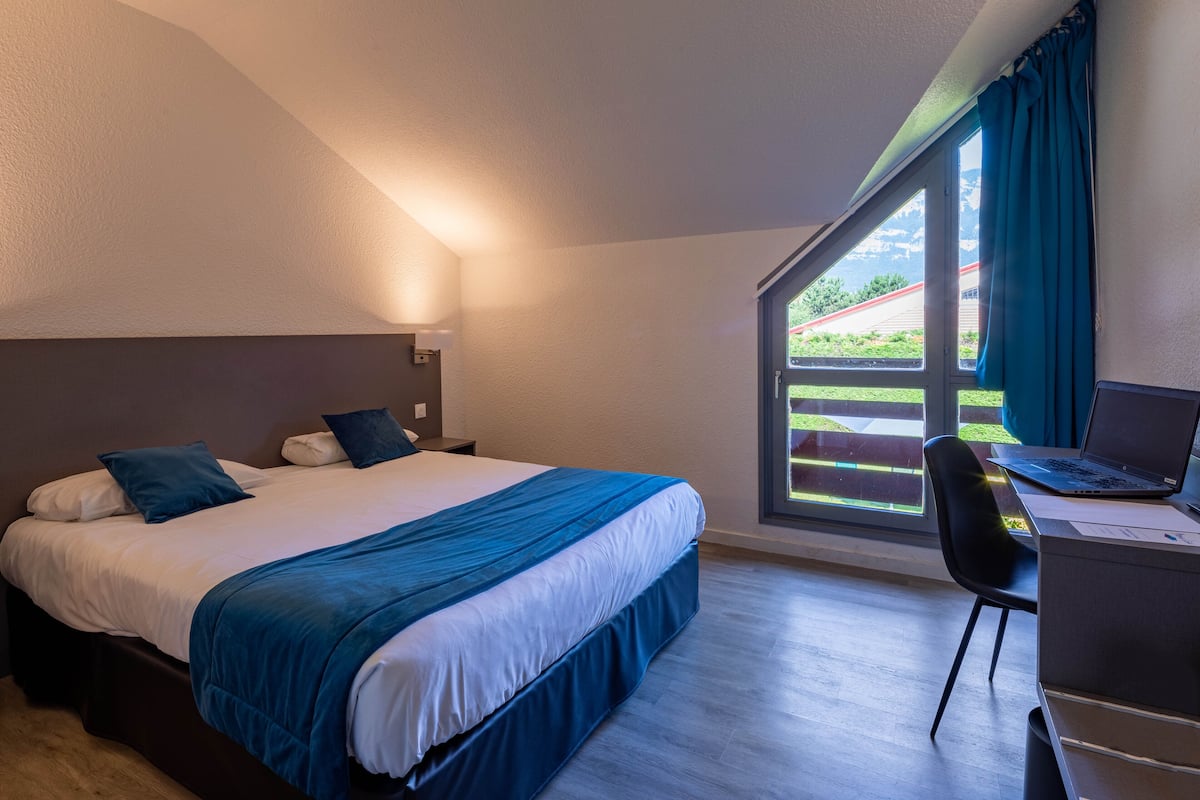 A bedroom with a bed, a desk with a laptop, and a window with curtains.
