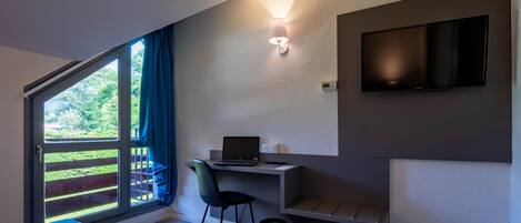 A modern bedroom with a large window, a desk with a laptop, a chair, and a TV mounted on the wall.