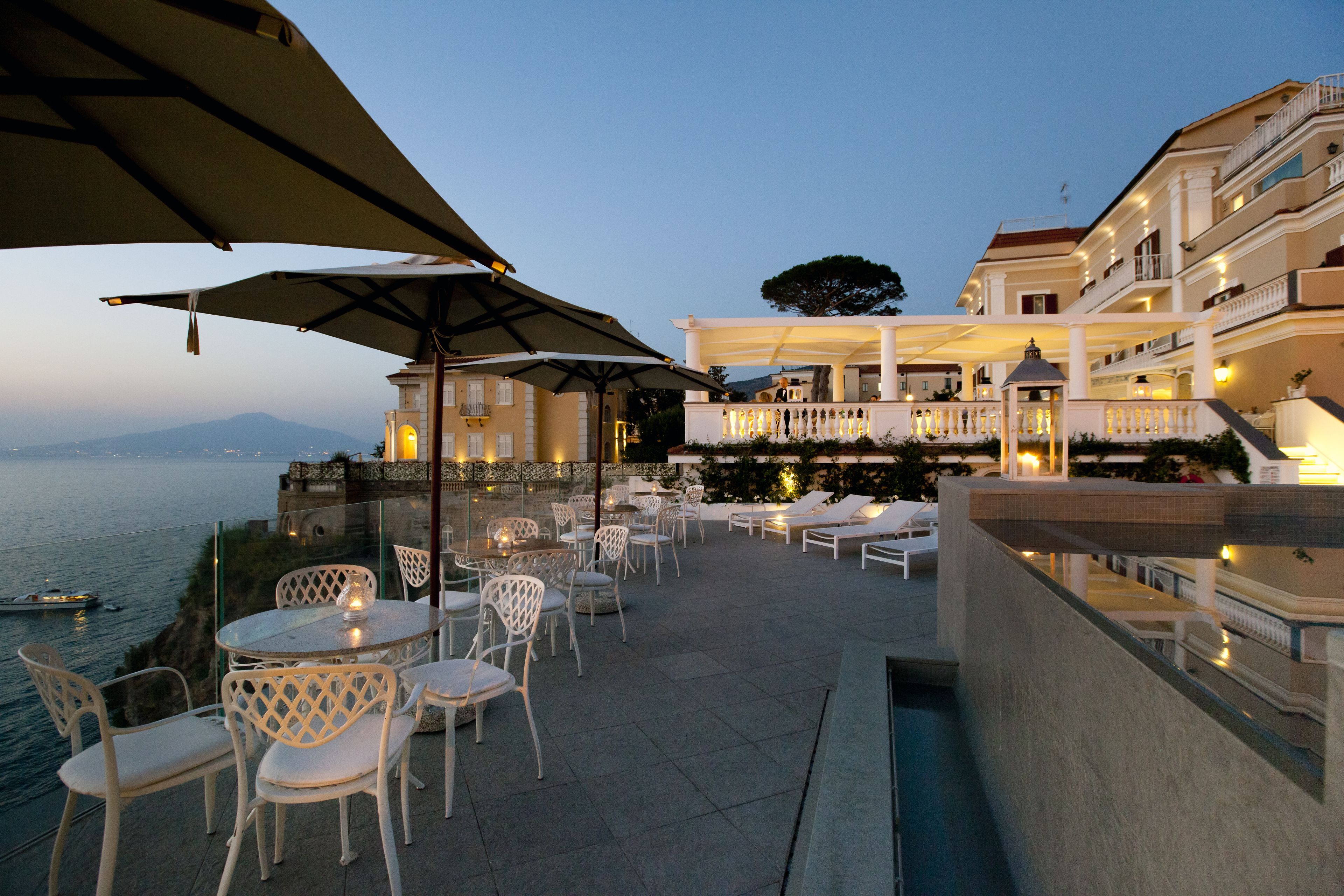 An outdoor terrace with tables and chairs, overlooking a body of water and a building with balconies.