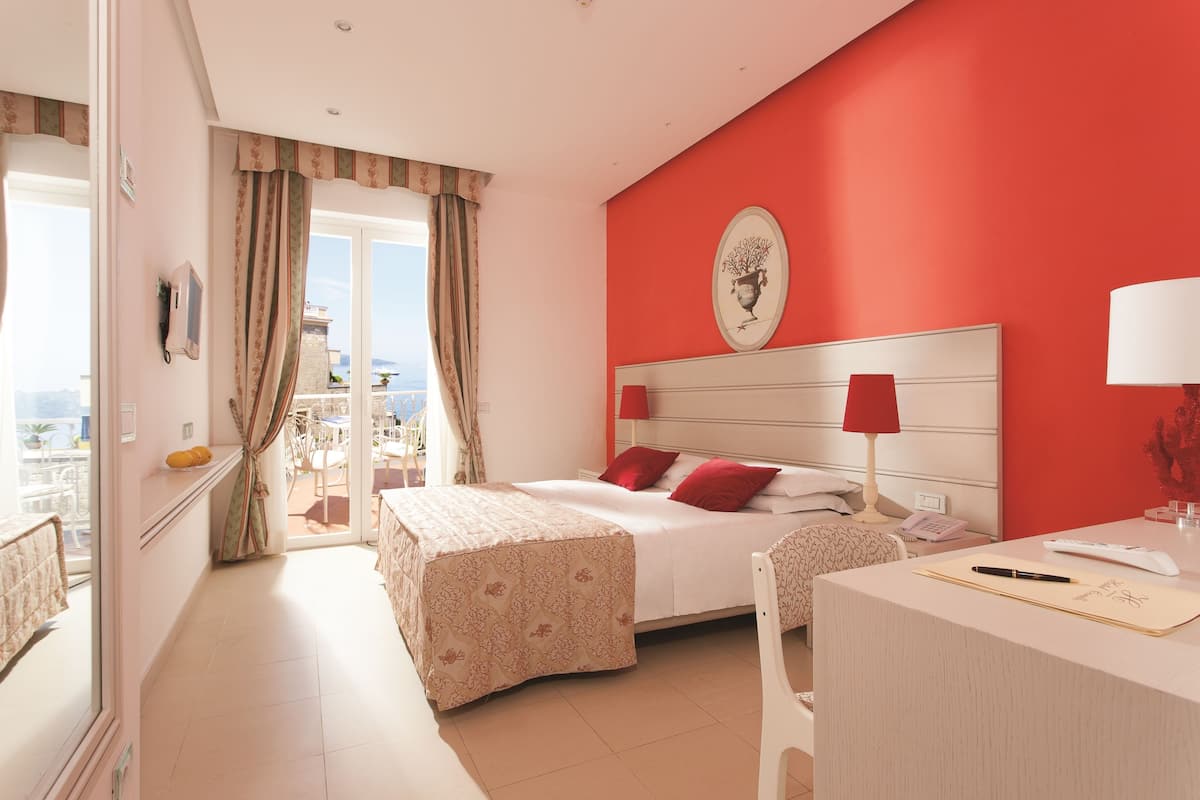 A hotel room with a bed, a desk, a chair, a lamp, and a view of the outdoors.