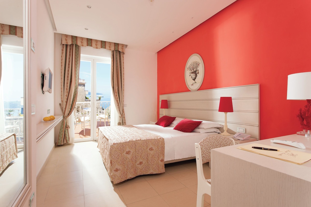 A hotel room with a bed, a desk, a chair, a lamp, and a view of the outside.