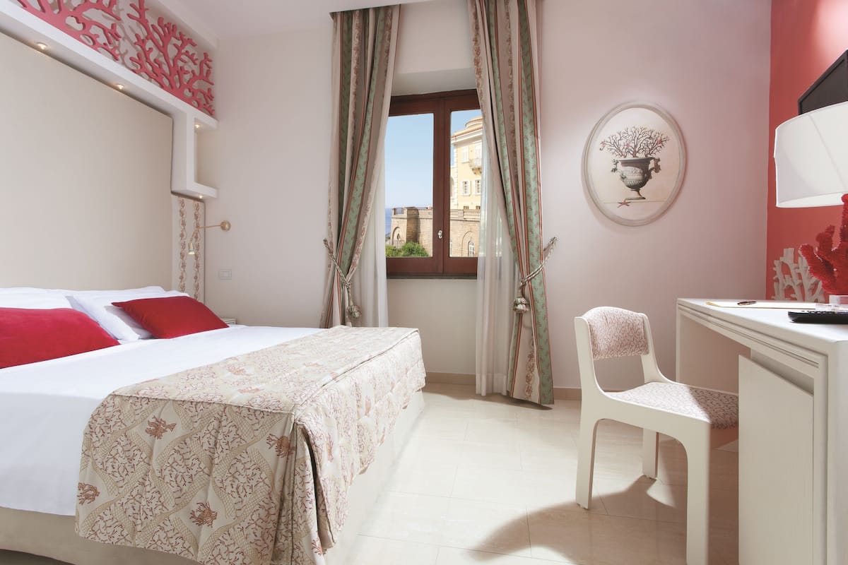 A hotel room with a bed, a desk, a chair, a window with curtains, and a wall decoration.