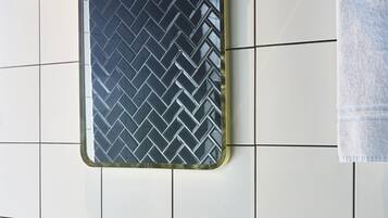 A bathroom with a white tiled wall, a black herringbone-patterned mirror, a white towel, a sink with a chrome faucet, and a small bottle of shampoo.