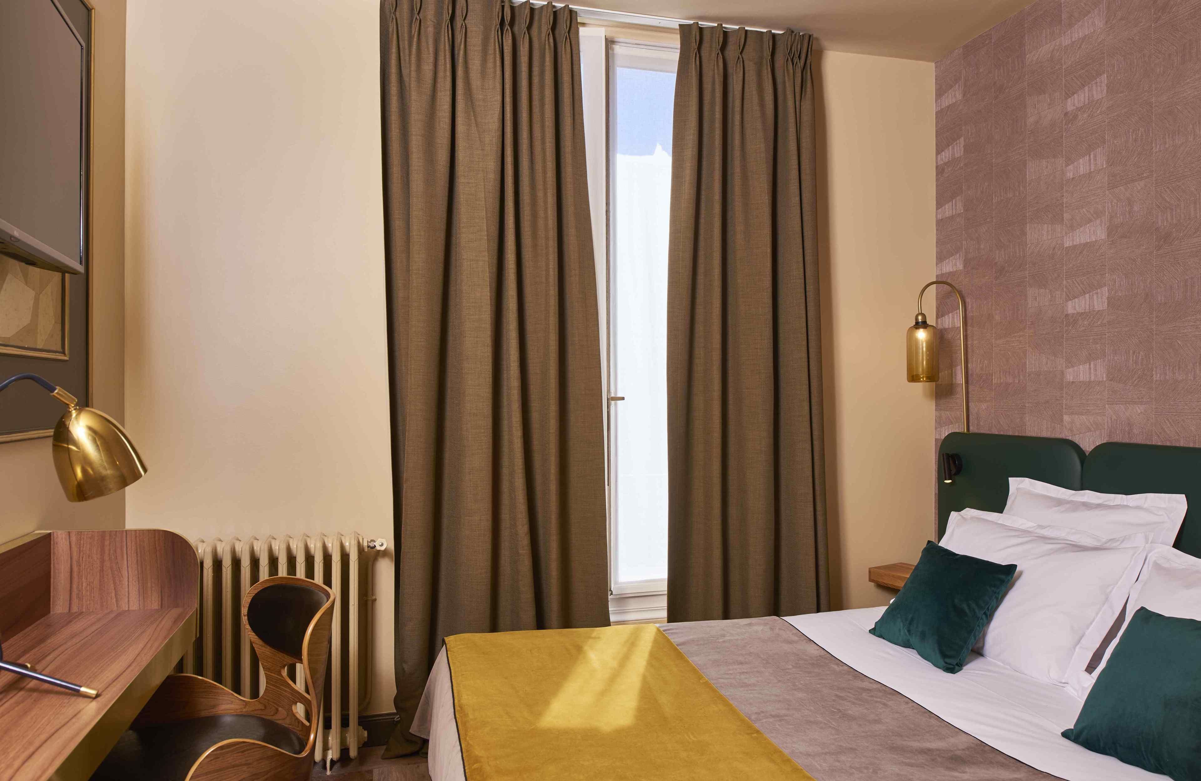 Habitació d'hotel amb llit, escriptori, cadira, làmpada i finestra amb cortines.