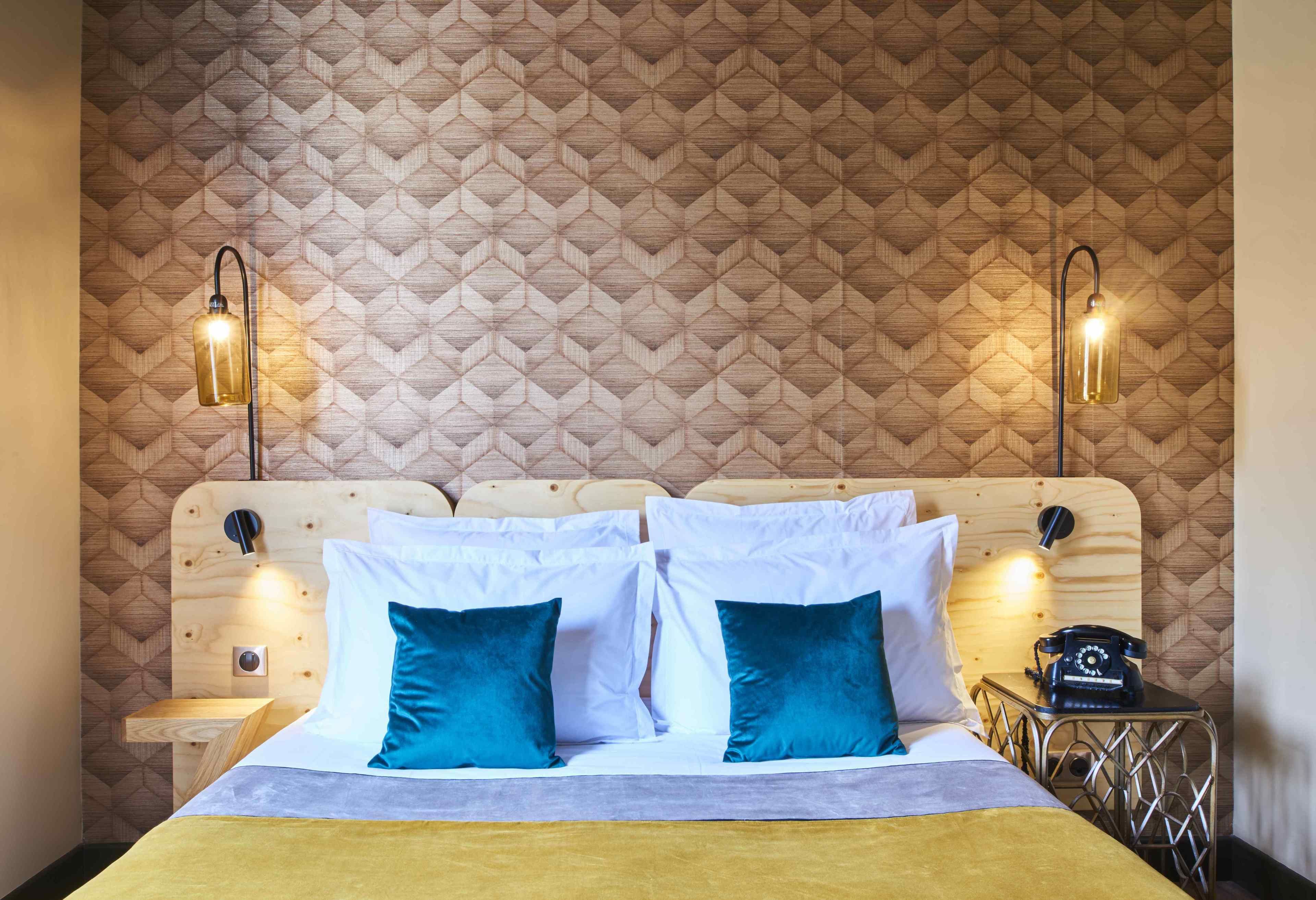 Une chambre d’hôtel avec une tête de lit en bois, deux lampes de chevet, un lit avec des oreillers blancs et turquoise, et un téléphone d’époque posé sur une table de chevet en métal.