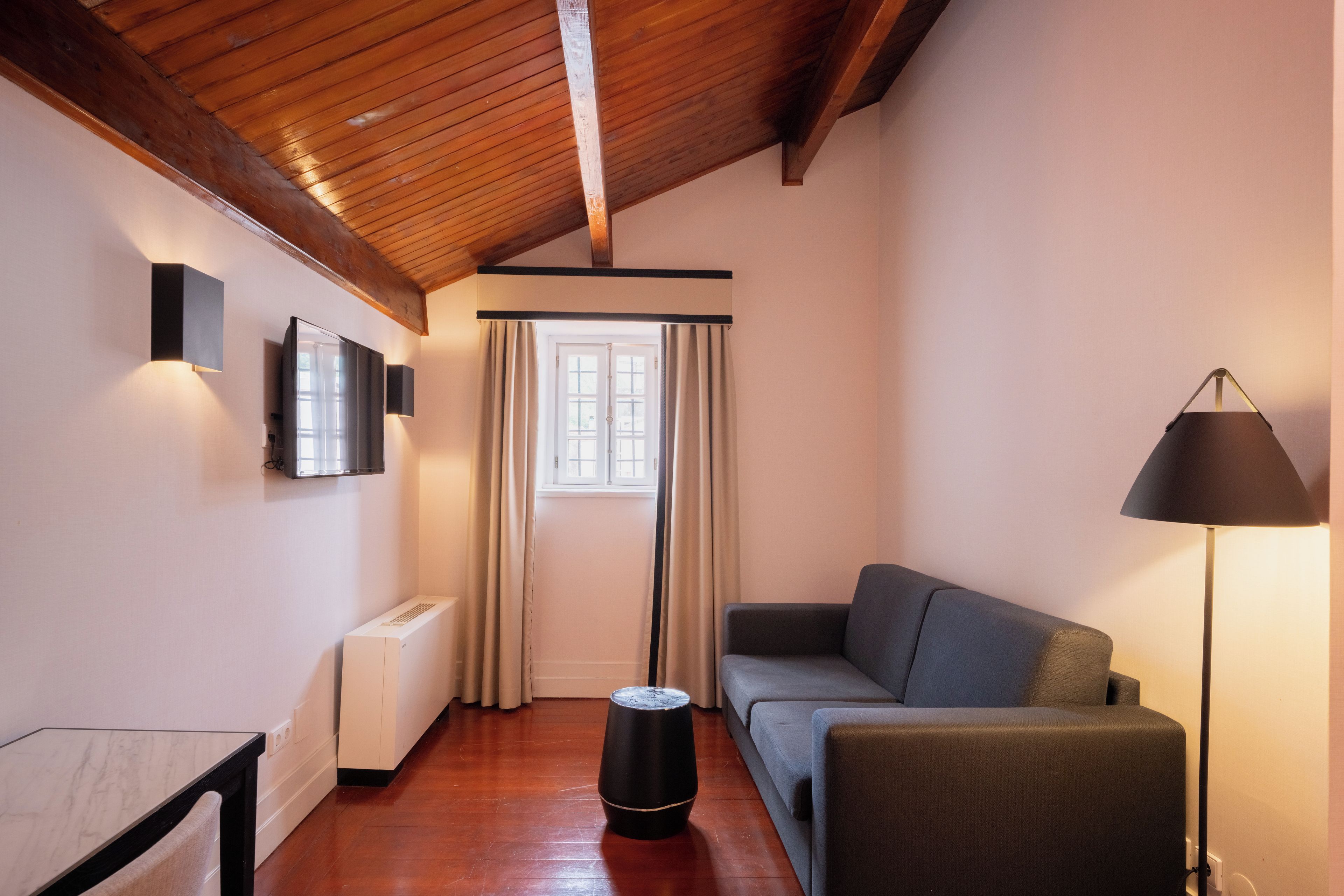 Una habitación con techo de madera, un sofá, una lámpara de pie, un radiador y una ventana con cortinas.