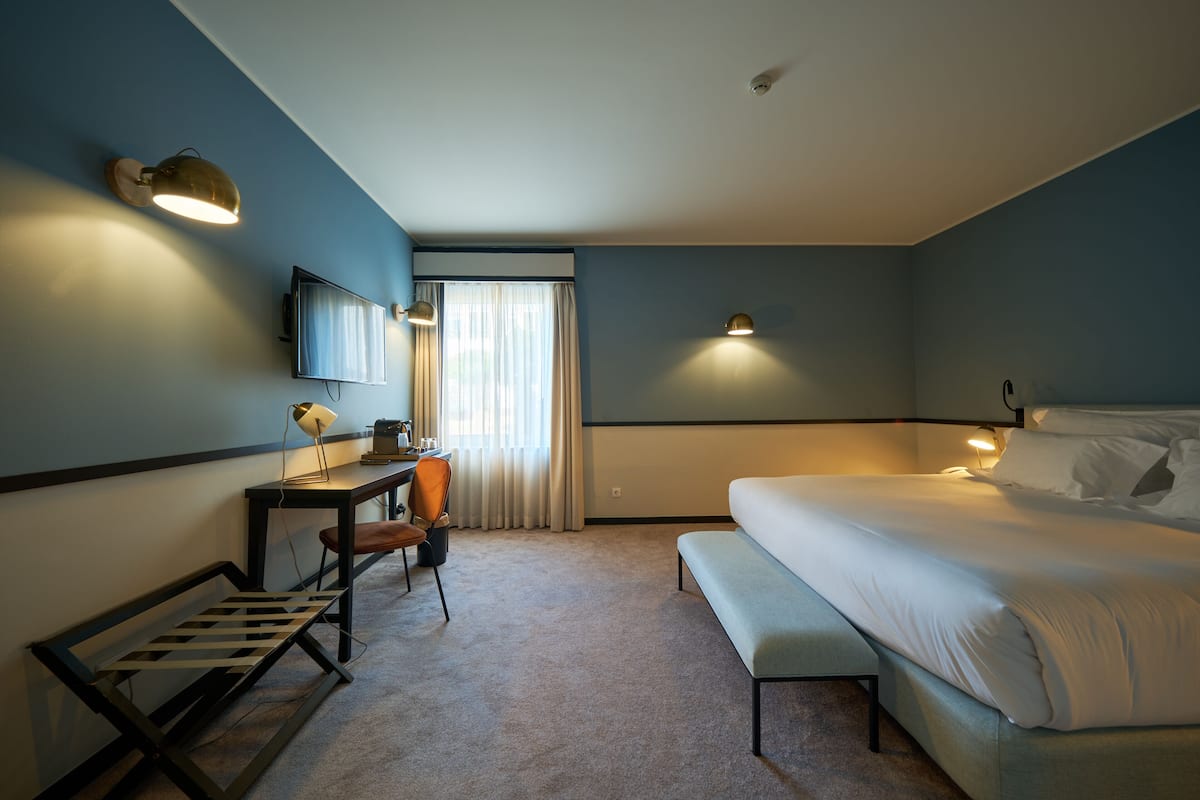 A hotel room with a bed, a desk with a chair, a bench, a TV, and two wall-mounted lamps.