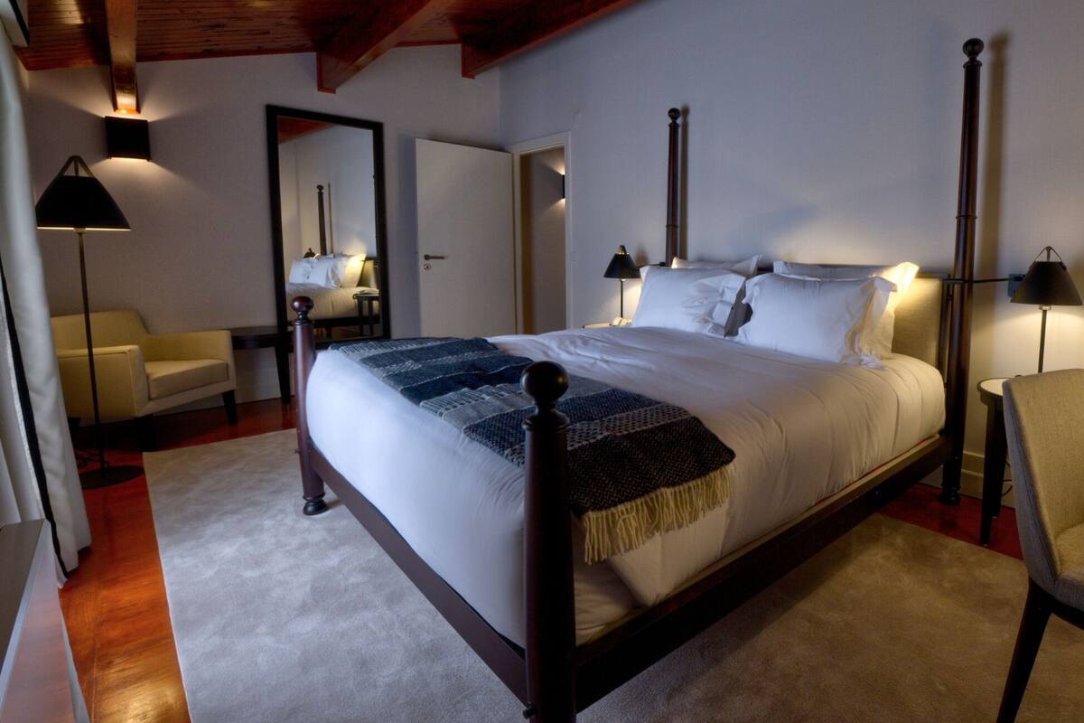 A neatly arranged bedroom with a large bed, bedside lamps, a chair, and a desk with a remote control.
