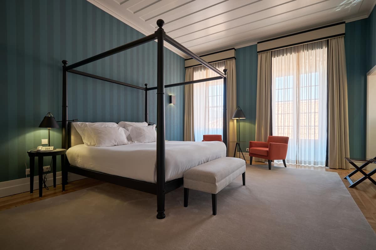 A four-poster bed with a matching bench, a red armchair, and a small table with a lamp.