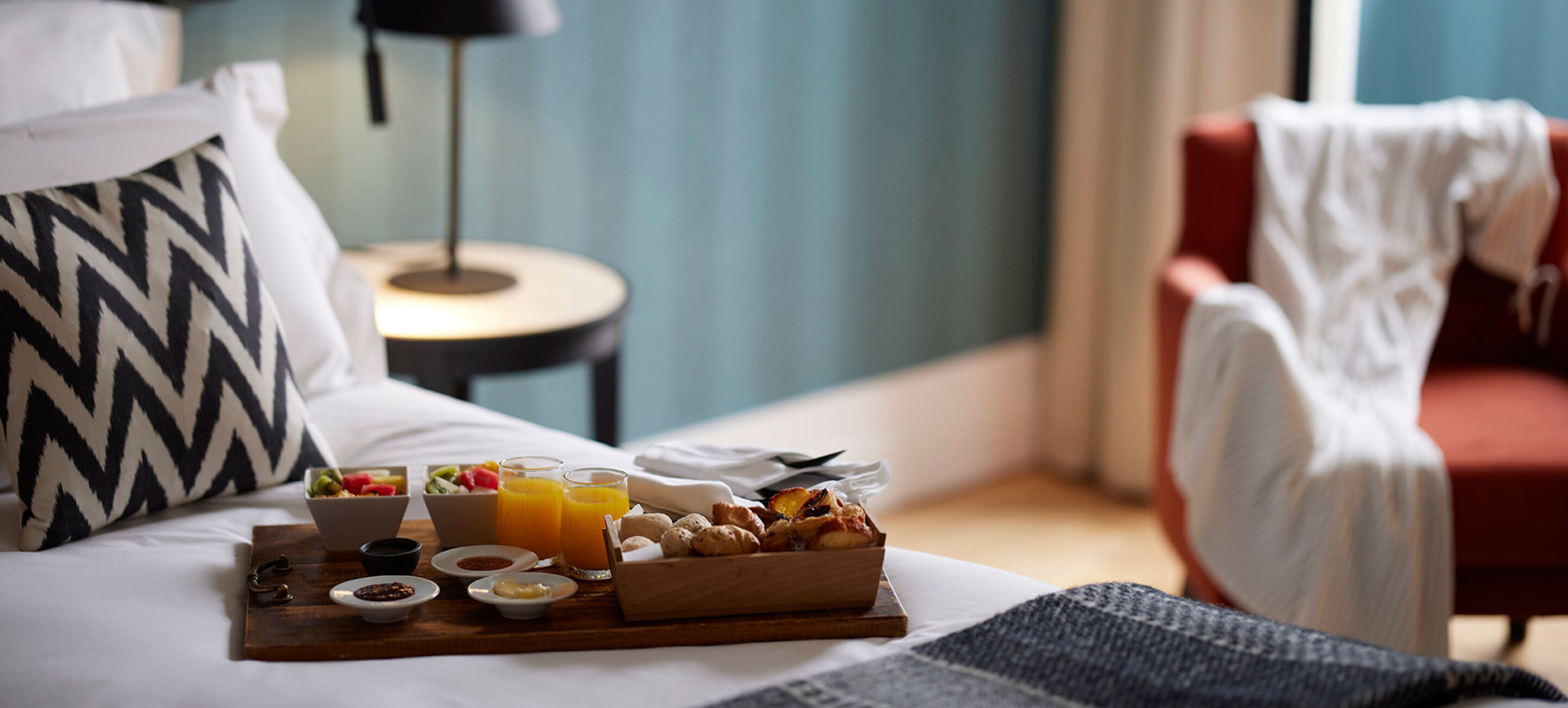 A neatly arranged breakfast tray on a bed with a pillow, a small table, and a chair in a room with a blue wall and a lamp.