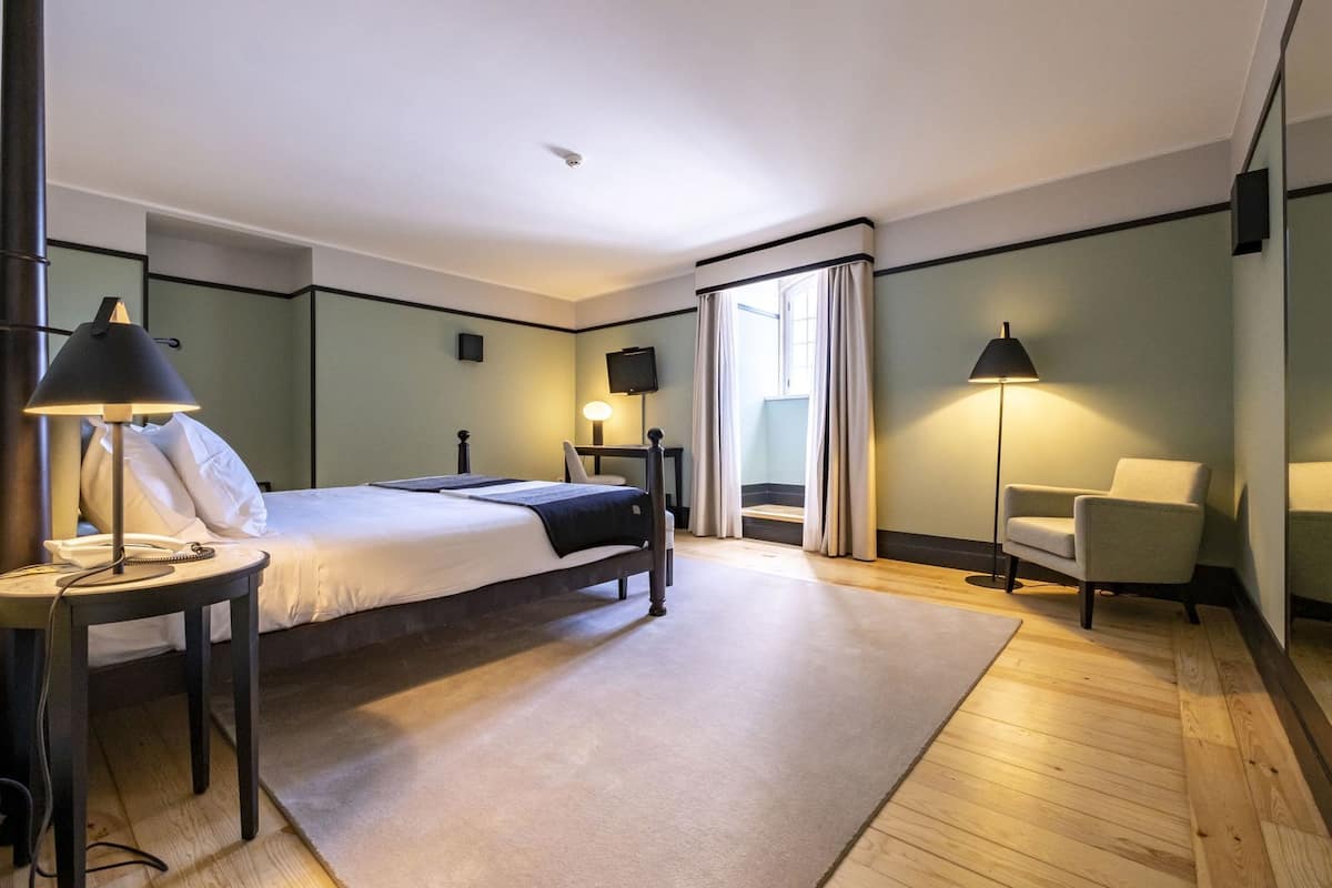 A hotel room with a bed, bedside tables, a chair, a TV, and a window with curtains.