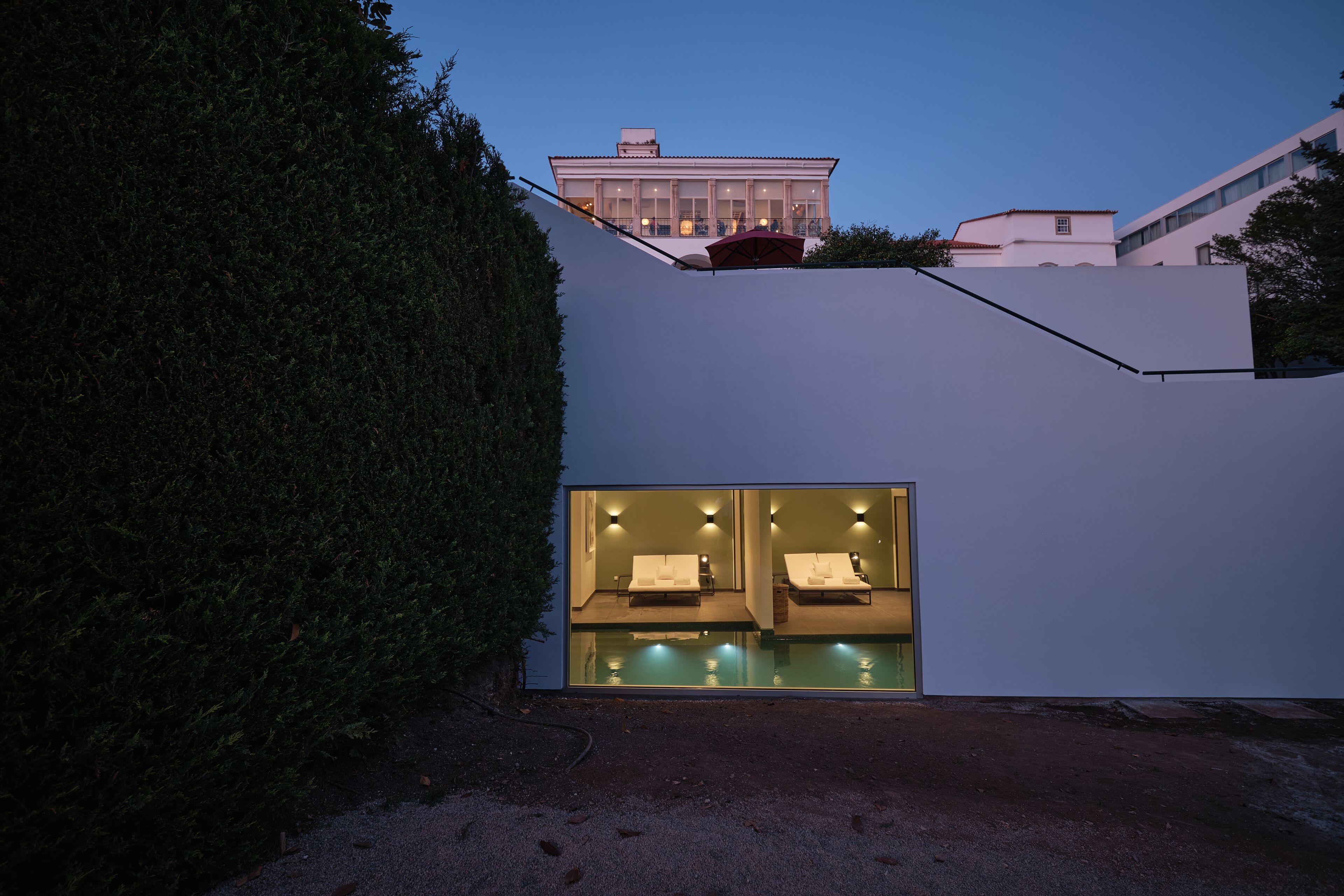 A modern building with a glass facade, a pool area with two loungers, and a well-maintained hedge.