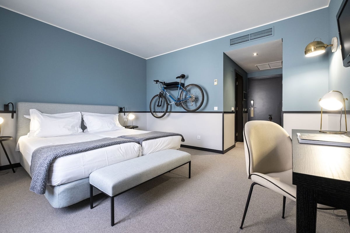 A modern hotel room with a bed, a desk, a chair, a bicycle, and a lamp.