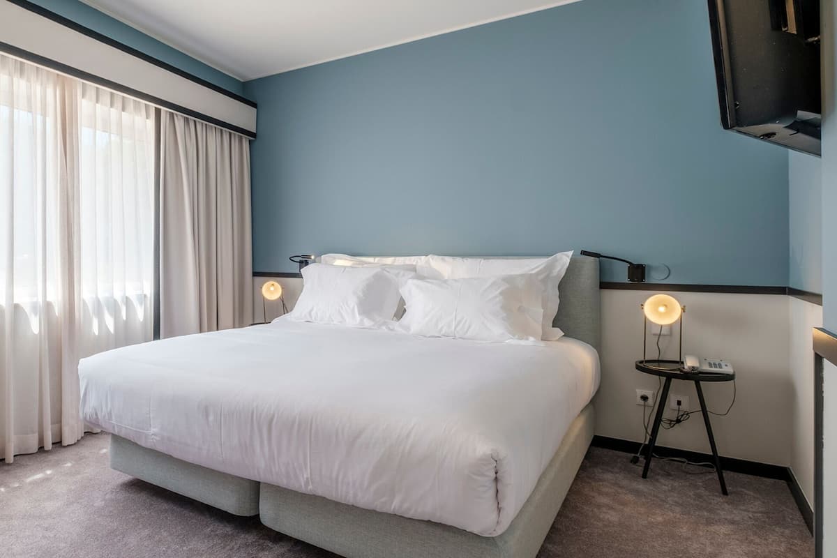 A hotel room with a bed, curtains, a bedside table with a lamp, and a wall-mounted TV.