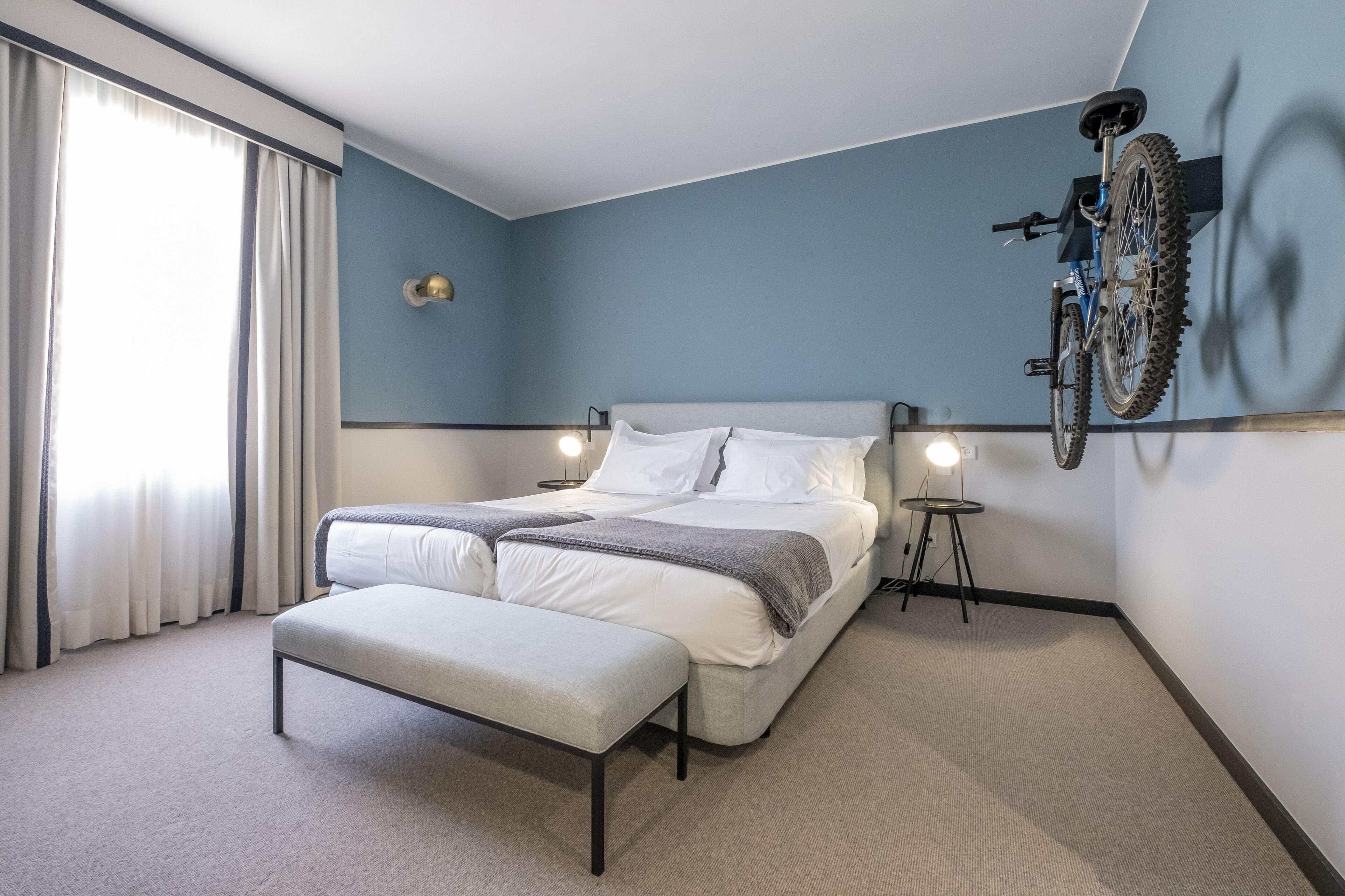 Et moderne hotelværelse med en stor seng, en bænk, en cykel monteret på væggen og et lille bord med en lampe.