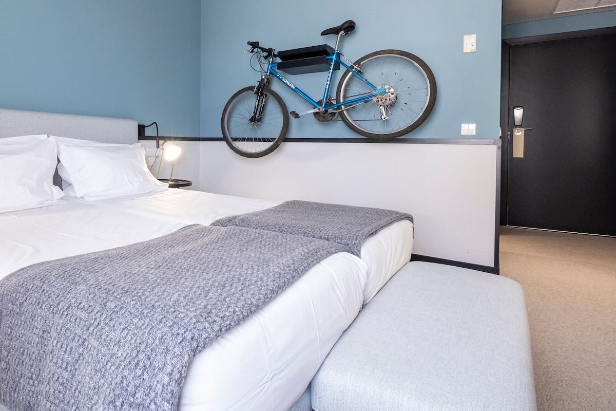 A neatly made bed with white linens and a grey blanket, a wall-mounted bicycle, and a bedside lamp.