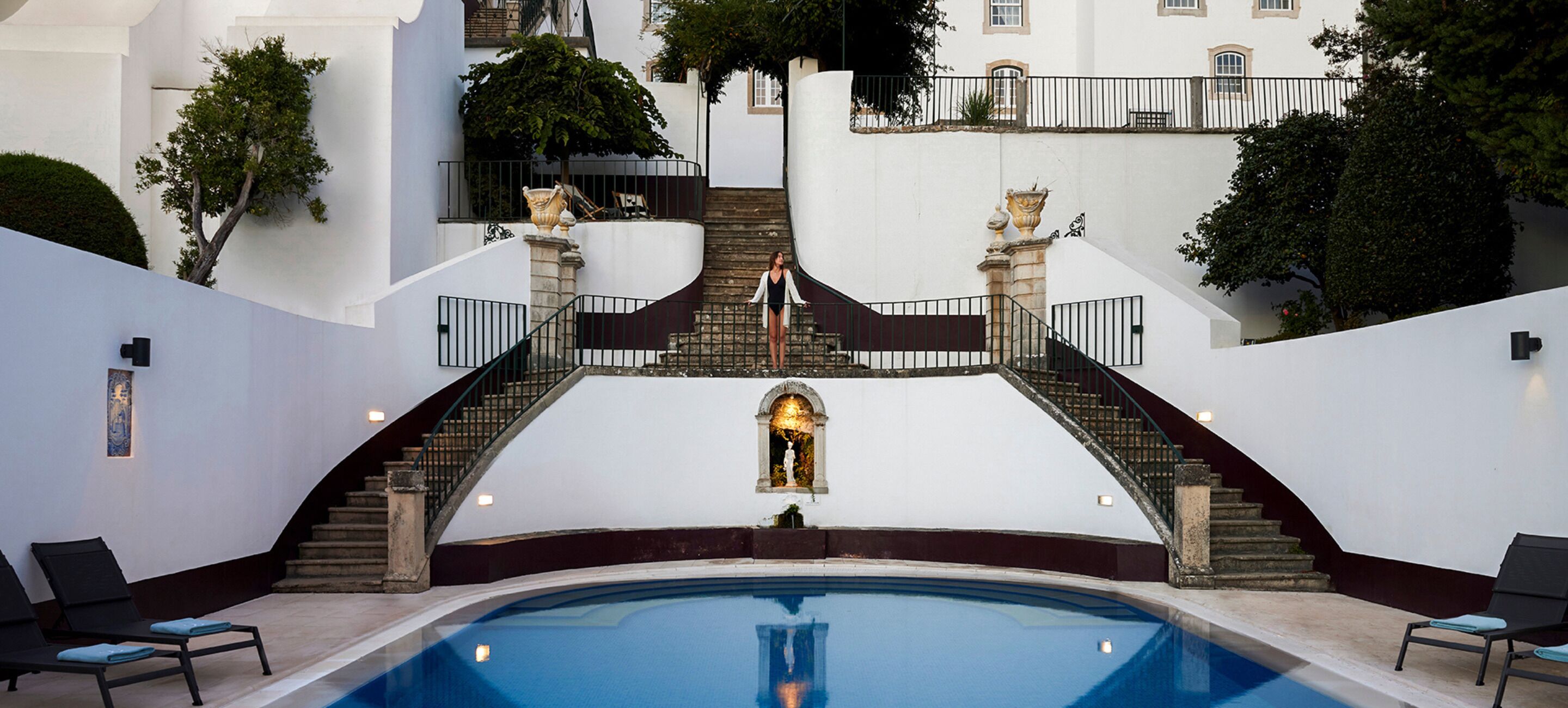 A pool area with a central staircase, lounge chairs, and a fountain.