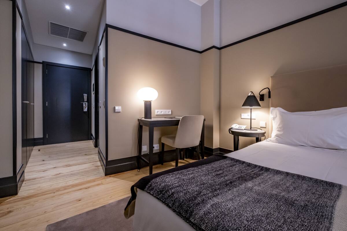 A hotel room with a bed, a desk with a chair, a lamp, and a phone on a side table.