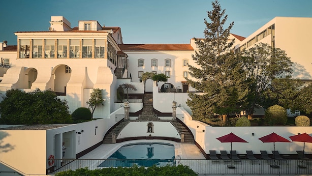 A large building with a central staircase leading to a pool area, surrounded by trees and a clear sky.