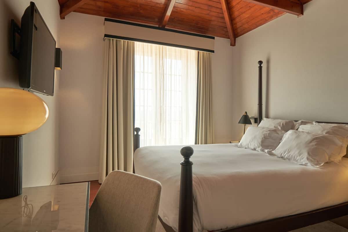 A hotel room with a bed, a chair, a desk, a lamp, a telephone, and a window with curtains.