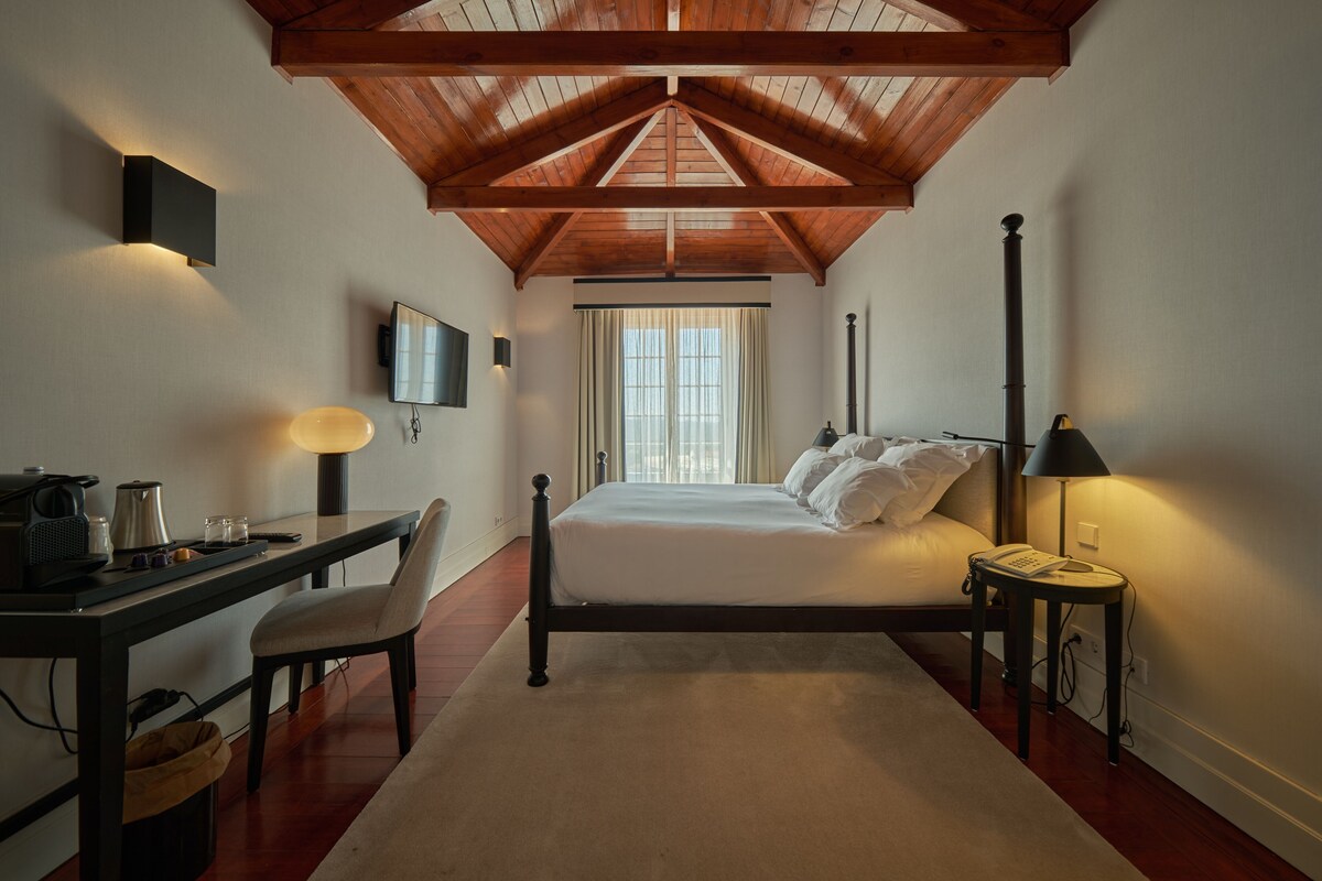 A bedroom with a wooden ceiling, a bed, a desk with a chair, and a nightstand with a lamp.