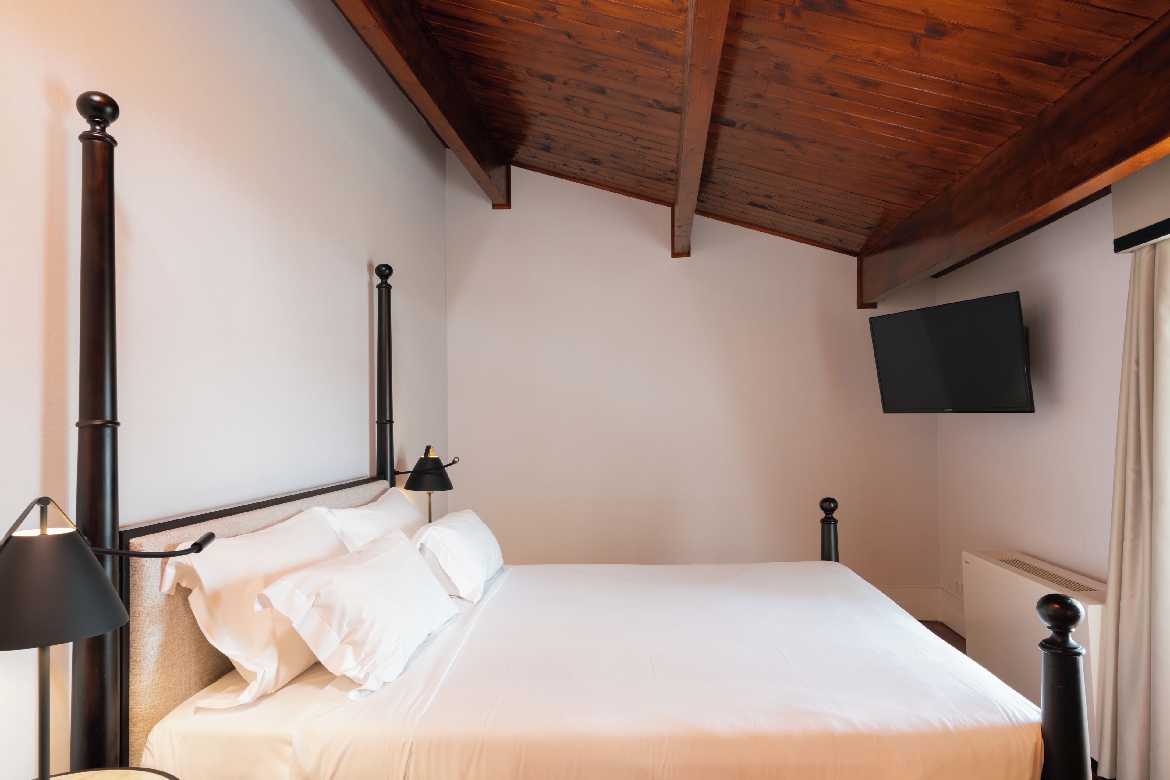 Un dormitorio con cama, televisor montado en la pared y ventana con cortinas.