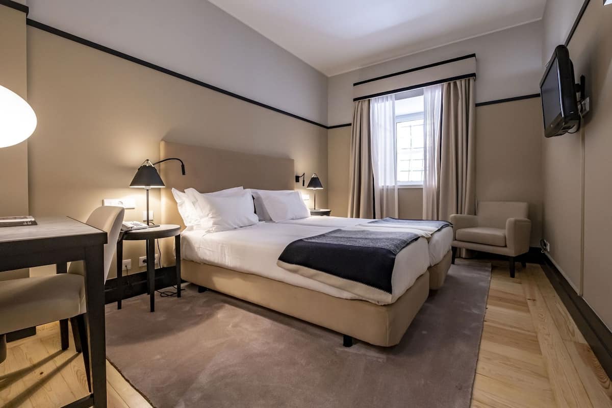 A hotel room with a bed, desk, chair, lamp, and a TV mounted on the wall.