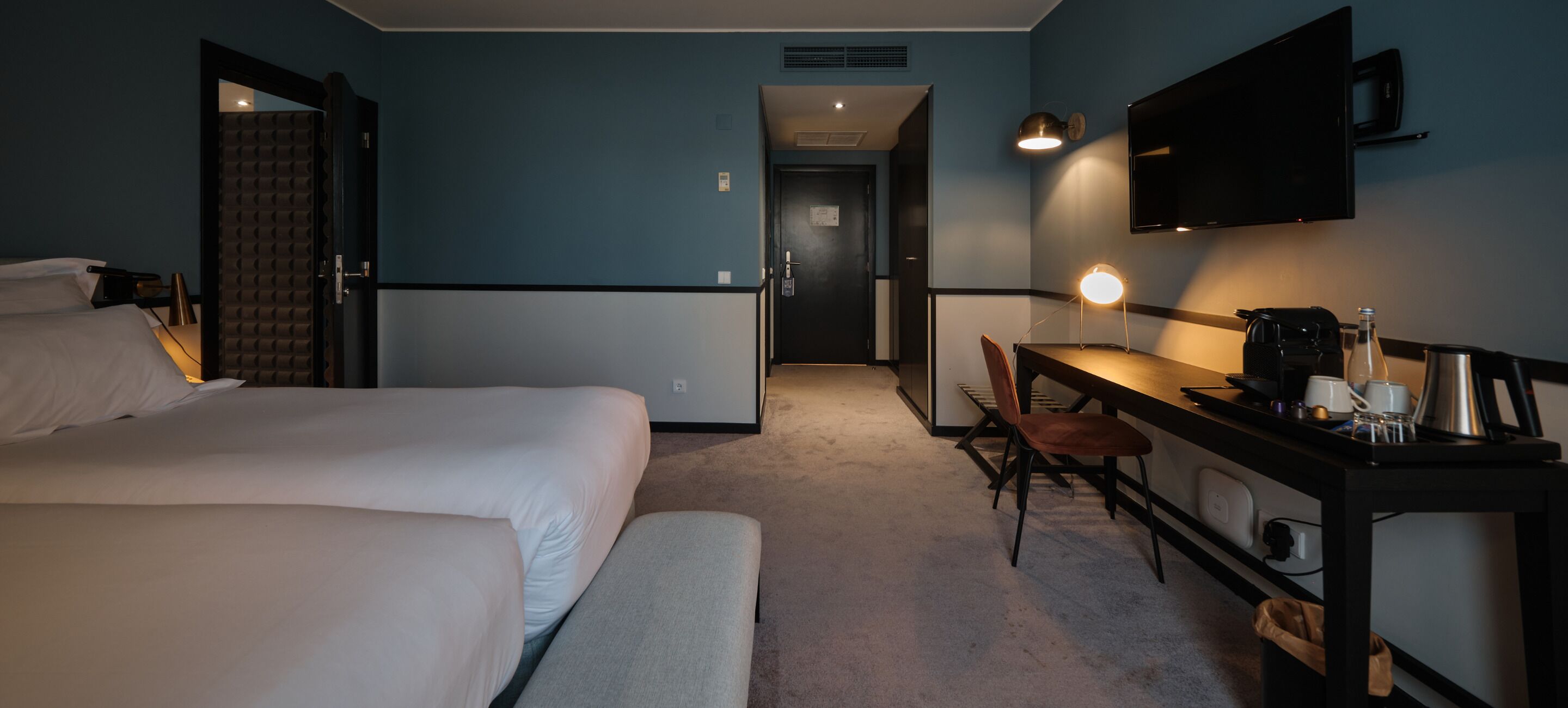Camera d'albergo con due letti, una scrivania con sedia, TV a parete e macchina per il caffè.