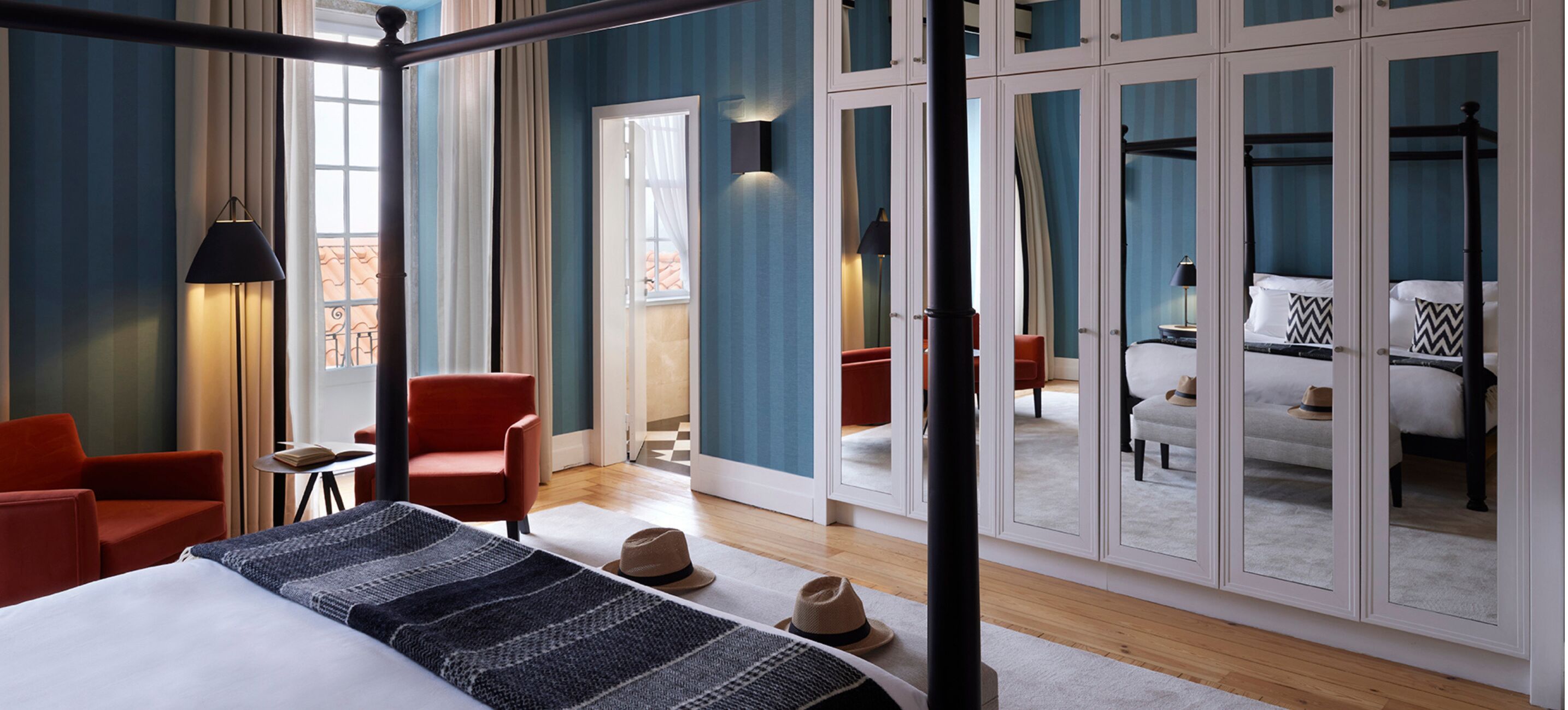 Habitación de hotel con una cama, un sillón rojo, una mesita con un libro y un armario grande con puertas de espejo.