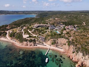 Vista aérea de un complejo turístico costero con un gran edificio, una piscina y un muelle que se extiende sobre aguas turquesas cristalinas.