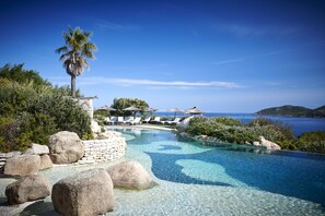 Piscina azul clara con sillas de descanso y una palmera, rodeada de rocas y follaje.