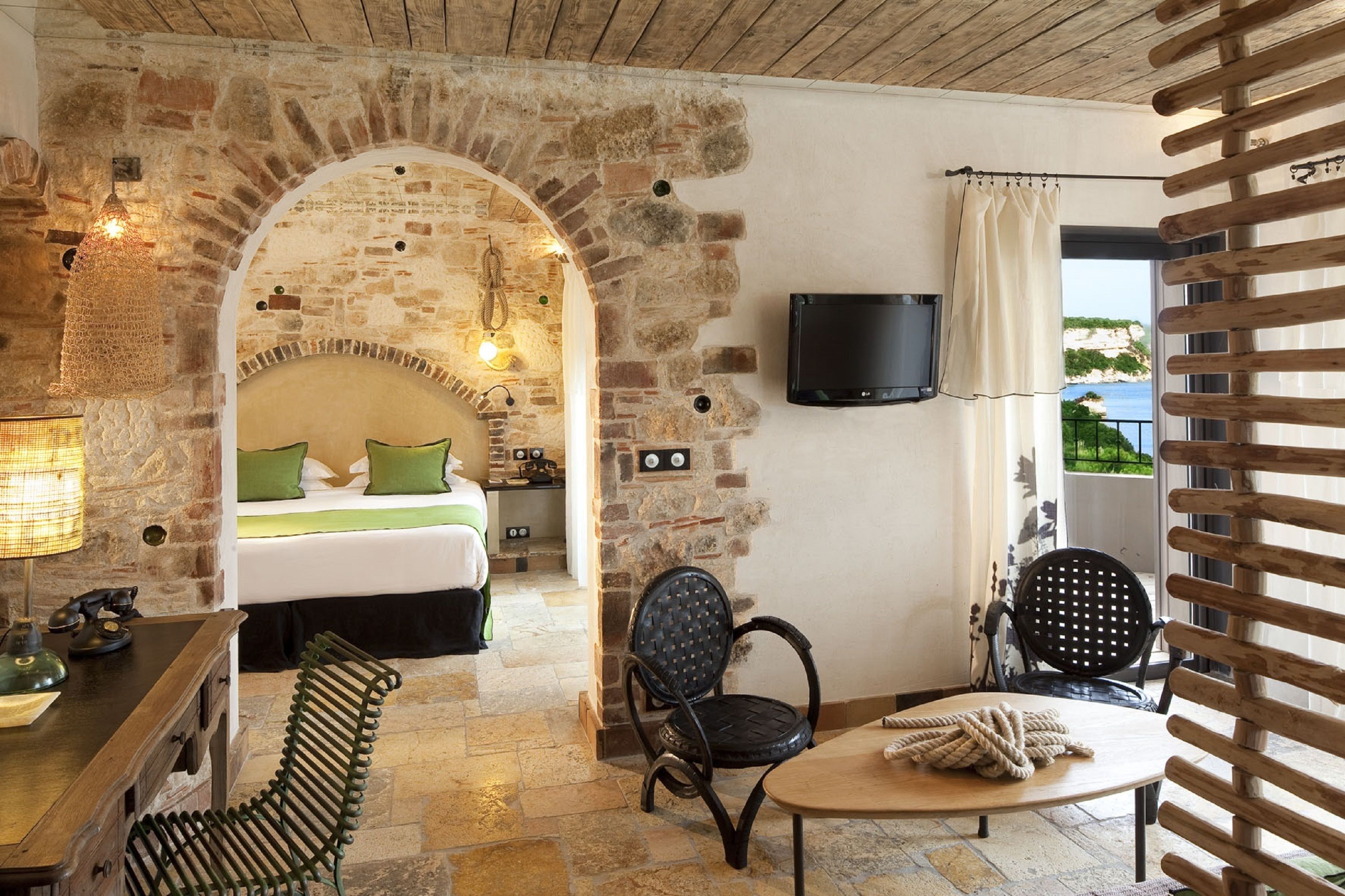 A cozy room with a stone fireplace, a bed with green pillows, a TV mounted on the wall, and a view of the sea through a window.