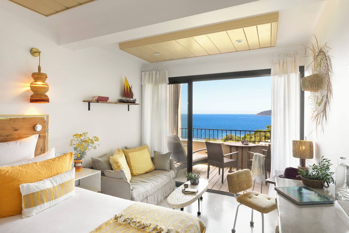 Un dormitorio amplio con una cama grande, un balcón con vistas al mar y una zona de comedor con mobiliario de mimbre.